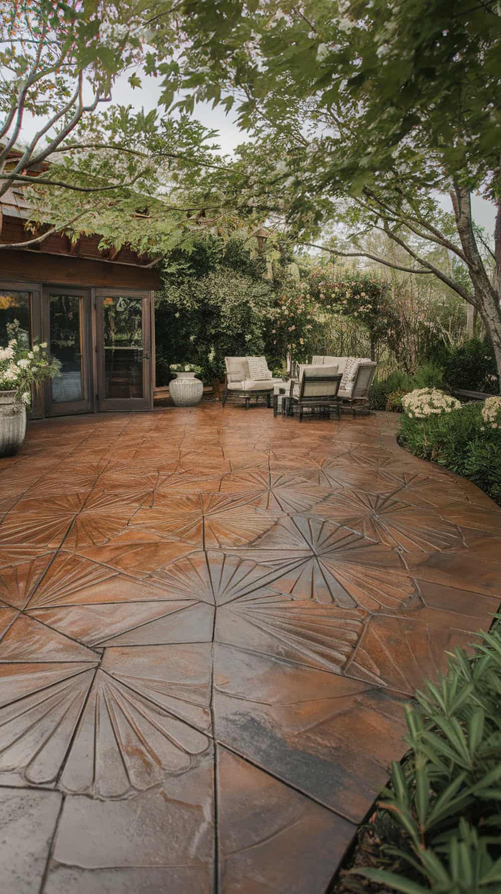 A beautifully designed stamped concrete patio with a fan pattern in rich brown tones, surrounded by greenery and outdoor furniture.