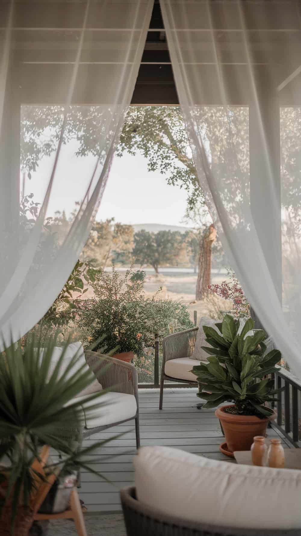 A cozy porch with sheer white curtains gently blowing in the breeze, surrounded by plants and comfortable seating.