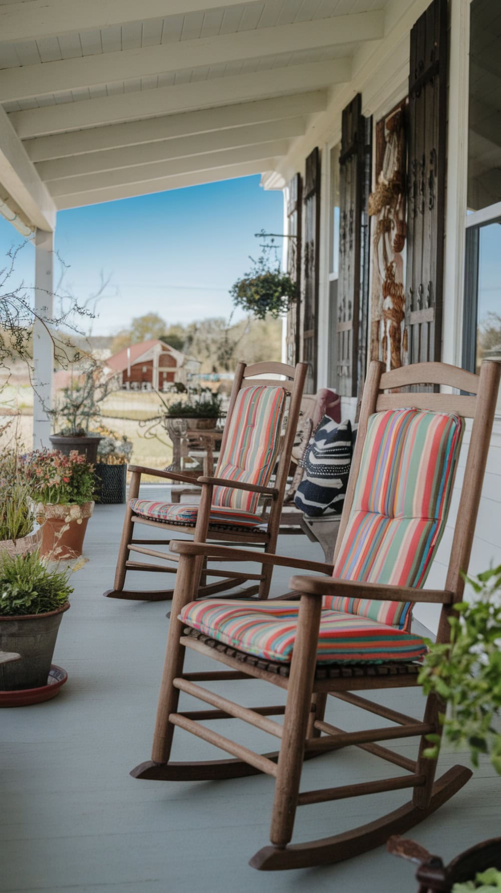 Two wooden rocking chairs with striped cushions on a farmhouse porch, surrounded by potted plants.